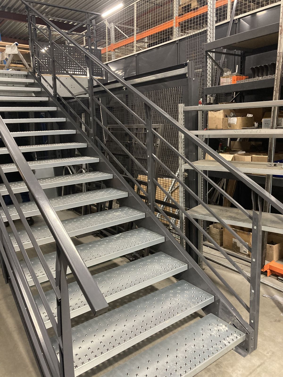 Verzinkte Gitterrost-Treppe mit schwarzen Wangen an einer Lagerbühne neben einem Palettenregal